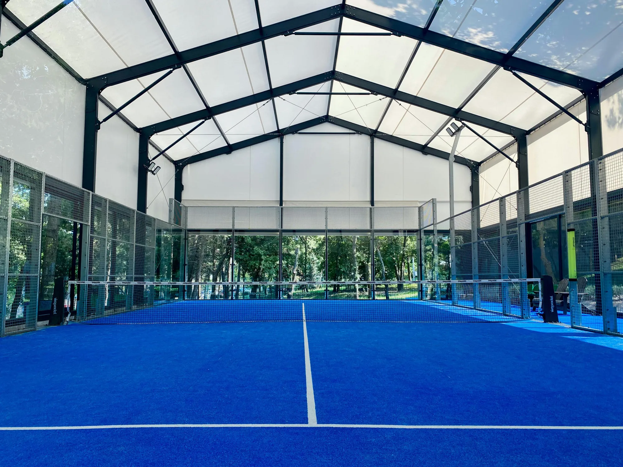 Indoor padel court with a blue surface, glass walls and a steel roof structure